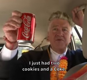 David Lynch sitting in a car, wearing a two-piece suit and a lei. He's holding up a can of Coca Cola, proudly declaring "I just had two cookies and a Coke."