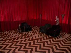 The Red Room. A red-tinted room with a rug featuring a zigzag pattern. Three chairs are arranged against the walls. One of the chairs has a statue of a naked woman behind them.